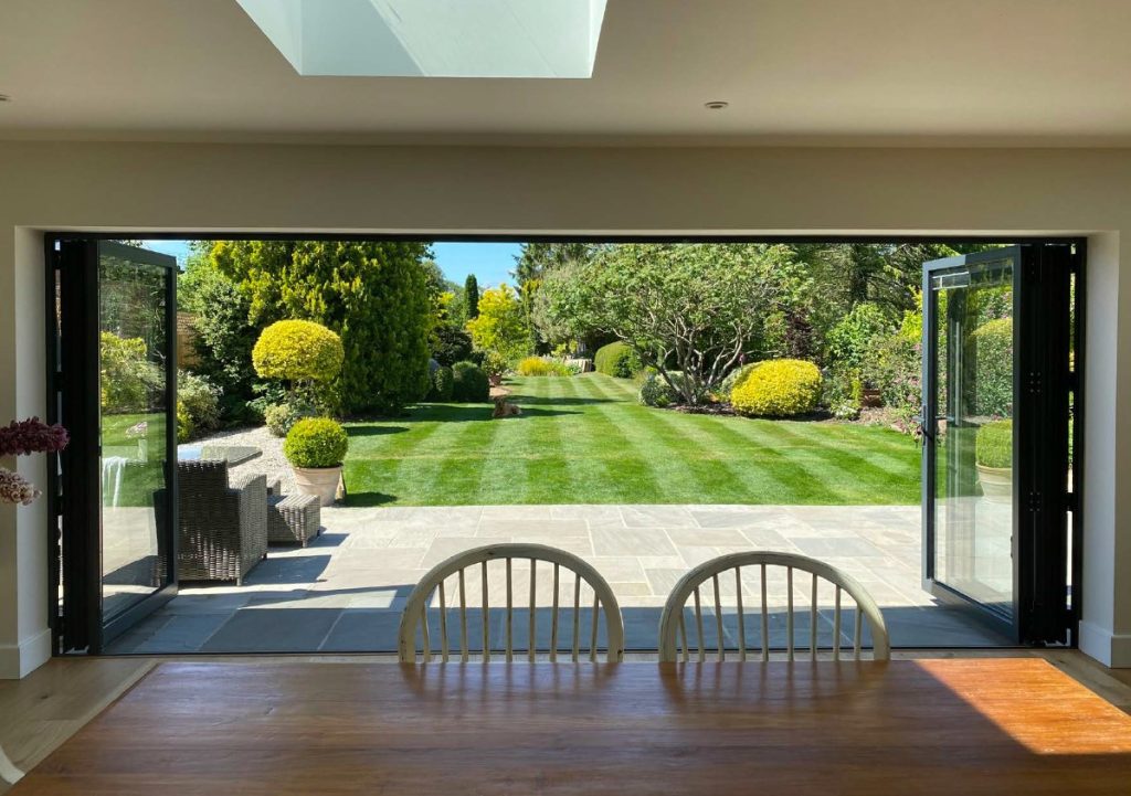 Dining area view of a lush garden with open glass doors under a skylight, wooden table in the foreground.