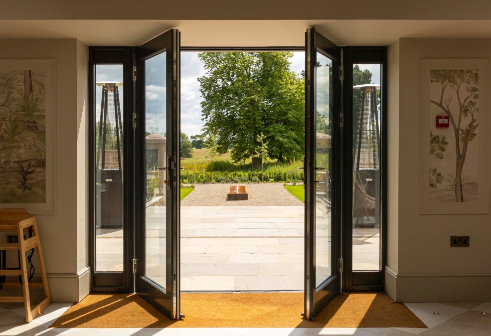 Wide open black bi-fold doors looking out onto a sunny patio and lush green landscape.